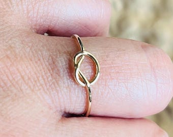 May include: A gold-coloured ring with a knot design. The ring is slender and delicate, with a polished finish. The knot is the central feature, creating a simple yet elegant aesthetic. The ring is shown on a finger.