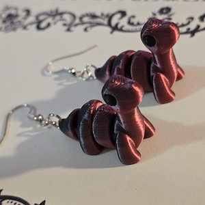 May include: A pair of burgundy dinosaur-shaped earrings. The earrings are 3D-printed with a textured finish and feature silver-colored ear wires. The dinosaur figures have a detailed, segmented body and a small head with a circular opening.