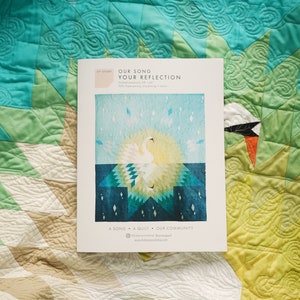May include: A white swan quilt pattern with a blue and green background. The pattern is called "Our Song Your Reflection" and is a song, a quilt, and a community.