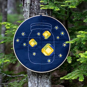Peut inclure: Une œuvre d'art en tissu bleu en forme de cercle représentant un bocal en verre avec des lucioles jaunes et vertes à l'intérieur. Le cercle est suspendu à un tronc d'arbre dans un environnement forestier.