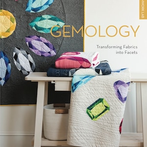 以下が含まれることがあります： アンドレア・ツァン・ジャクソン著「ジェモロジー：ファブリックをファセットに変える」というタイトルの本。本の表紙には、宝石のような幾何学模様のカラフルなキルトが描かれています。