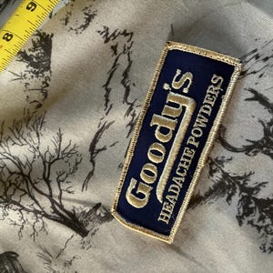 May include: A rectangular navy blue patch with gold lettering that reads "Goody's Headache Powders." The patch has a gold border and is set against a patterned fabric. A yellow measuring tape is in the upper left corner.