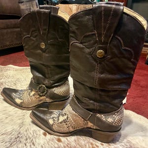Op de afbeelding: Een paar bruine leren cowboylaarzen met slangenleeraccenten. De laarzen hebben een puntige neus, een gespdetail en een gestapelde hak. De schacht van de laarzen is hoog en het leer lijkt versleten.