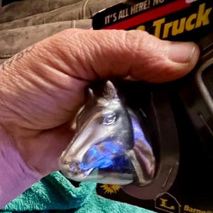 May include: A chrome horse head truck emblem. The emblem is held in a person's hand. The packaging reads "Truck". The horse head is a detailed sculpture with a blue light reflecting off of it.