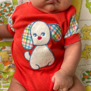 Puede incluir: Un beb&eacute; con un mono rojo con un cachorro blanco bordado en la parte delantera. El cachorro tiene orejas a cuadros y una nariz roja.