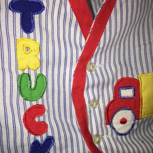 May include: A white and blue striped shirt with a red trim and a red, yellow, and blue embroidered truck on the right side. The word "TRUCK" is embroidered in colorful letters on the left side of the shirt.