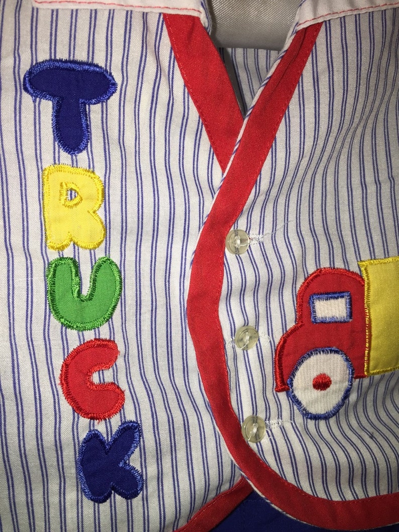 May include: A white and blue striped shirt with a red trim and a red, yellow, and blue embroidered truck on the right side. The word "TRUCK" is embroidered in colorful letters on the left side of the shirt.