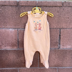 K&ouml;nnte beinhalten: Ein hellgelber, &auml;rmelloser Baby-Strampler mit einem Aufdruck von zwei Giraffen auf einem H&uuml;gel. Der Strampler hat einen Knopfverschluss an der Schulter.