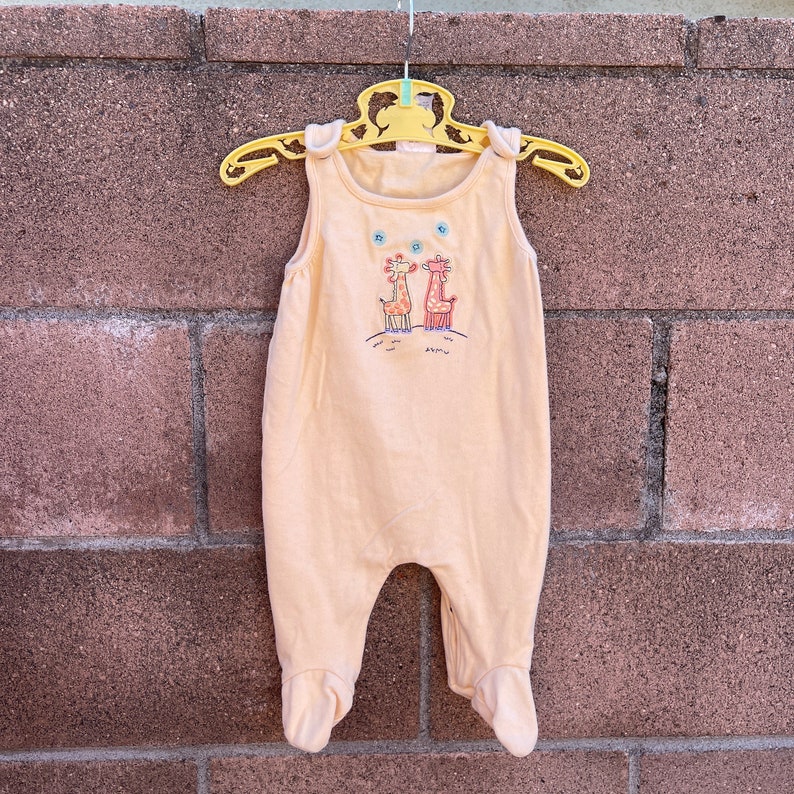 K&ouml;nnte beinhalten: Ein hellgelber, &auml;rmelloser Baby-Strampler mit einem Aufdruck von zwei Giraffen auf einem H&uuml;gel. Der Strampler hat einen Knopfverschluss an der Schulter.