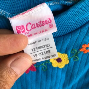 May include: A blue baby onesie with embroidered flowers. The tag reads "Carter's" "Made in U.S.A." "12 Months" "19-22 LBS." "Content & Care Over"