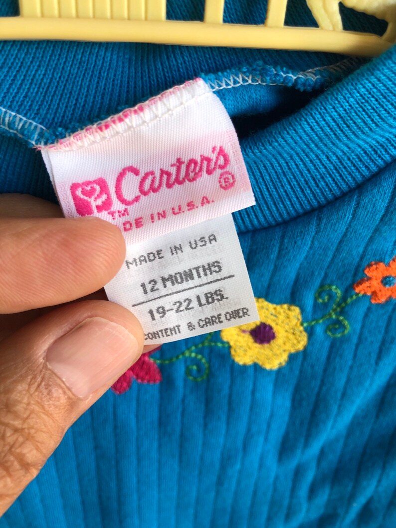 May include: A blue baby onesie with embroidered flowers. The tag reads "Carter's" "Made in U.S.A." "12 Months" "19-22 LBS." "Content & Care Over"