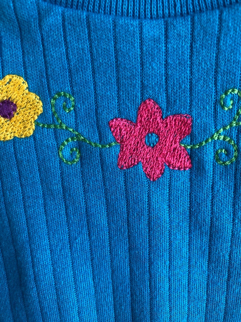 May include: A blue ribbed jumper with embroidered flowers in yellow, pink, and green.