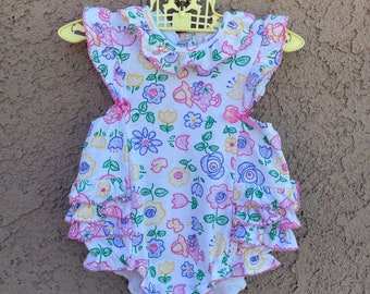 May include: A white baby onesie with a pink and blue floral print. The onesie has a ruffled trim and is sleeveless.