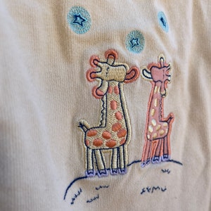 K&ouml;nnte beinhalten: Ein hellgelber Baby-Strampler mit einem gestickten Design aus zwei Giraffen, die zu blauen Sternen aufblicken. Die Giraffen sind braun und orange mit lila F&uuml;&szlig;en.