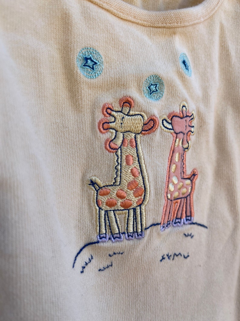 K&ouml;nnte beinhalten: Ein hellgelber Baby-Strampler mit einem gestickten Design aus zwei Giraffen, die zu blauen Sternen aufblicken. Die Giraffen sind braun und orange mit lila F&uuml;&szlig;en.