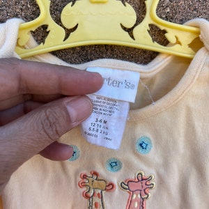 K&ouml;nnte beinhalten: Ein gelber Baby-Strampler mit Giraffen-Druck und blauen Sternen. Der Strampler ist aus 100% Baumwolle und tr&auml;gt das Label "Carter's". Das Gr&ouml;&szlig;enetikett zeigt "3-6 M", "12-18 lbs", "5,5-8 kg".