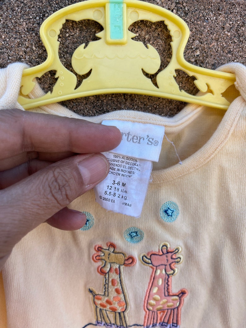 K&ouml;nnte beinhalten: Ein gelber Baby-Strampler mit Giraffen-Druck und blauen Sternen. Der Strampler ist aus 100% Baumwolle und tr&auml;gt das Label "Carter's". Das Gr&ouml;&szlig;enetikett zeigt "3-6 M", "12-18 lbs", "5,5-8 kg".