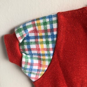 Puede incluir: Una camisa roja de manga corta con un patr&oacute;n de cuadros en las mangas. El patr&oacute;n de cuadros es en tonos de azul, verde, amarillo y rojo.