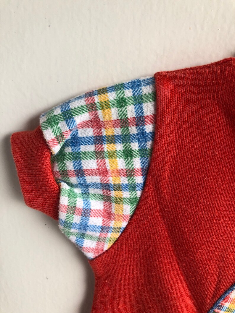 Puede incluir: Una camisa roja de manga corta con un patr&oacute;n de cuadros en las mangas. El patr&oacute;n de cuadros es en tonos de azul, verde, amarillo y rojo.