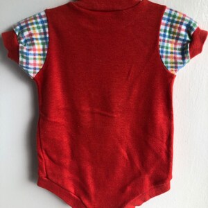 Puede incluir: Unos&iacute; rojo para beb&eacute; con cuello alto y mangas cortas. Las mangas tienen un patr&oacute;n de cuadros en azul, verde, amarillo y blanco.