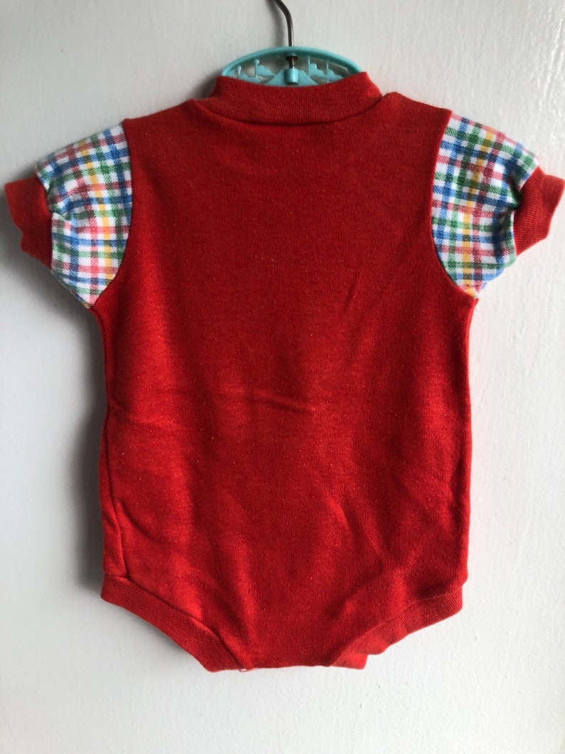 Puede incluir: Unos&iacute; rojo para beb&eacute; con cuello alto y mangas cortas. Las mangas tienen un patr&oacute;n de cuadros en azul, verde, amarillo y blanco.