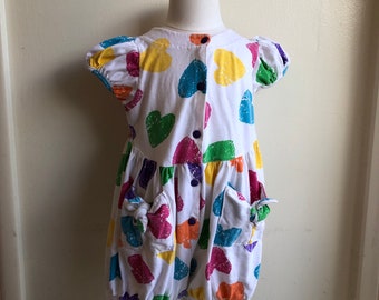 May include: A white dress with colorful heart print and pockets. The dress has a button closure down the front.