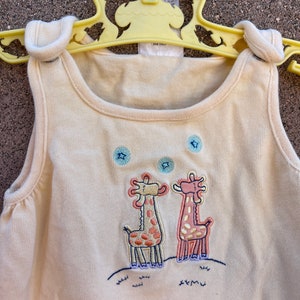 K&ouml;nnte beinhalten: Ein cremefarbener, &auml;rmelloser Strampler mit einem Motiv von zwei Giraffen, die auf einem grasbewachsenen H&uuml;gel mit Sternen am Himmel stehen. Die Giraffen sind in Rosa, Gelb und Braun gestickt.