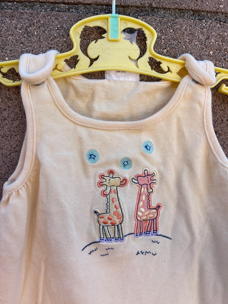 K&ouml;nnte beinhalten: Ein cremefarbener, &auml;rmelloser Strampler mit einem Motiv von zwei Giraffen, die auf einem grasbewachsenen H&uuml;gel mit Sternen am Himmel stehen. Die Giraffen sind in Rosa, Gelb und Braun gestickt.