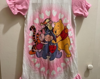 May include: A white nightgown with pink sleeves and a pink ruffled hem. The center features a graphic of Winnie the Pooh, Tigger, Piglet, and Eeyore hugging. Eeyore holds a red heart with the text UR Luvd, surrounded by a pink heart border.