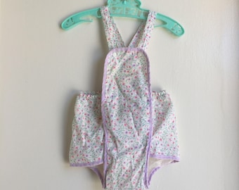 May include: A vintage baby romper with a floral pattern in pink and green on a white background, trimmed with lavender. The romper is hanging on a light green plastic hanger.