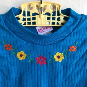 May include: A blue ribbed knit shirt with a floral embroidery design around the neckline. The shirt has a tag that reads "Carter's" and "Made in U.S.A."