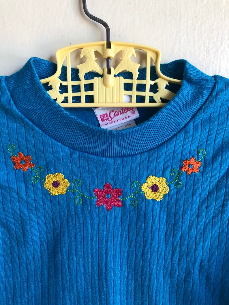 May include: A blue ribbed knit shirt with a floral embroidery design around the neckline. The shirt has a tag that reads "Carter's" and "Made in U.S.A."