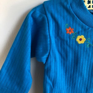 May include: A blue long-sleeved shirt with a ribbed texture and embroidered flowers in orange, yellow, and green.