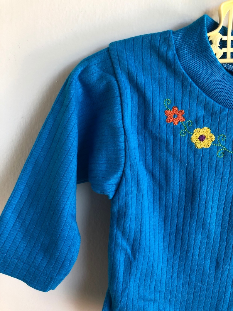 May include: A blue long-sleeved shirt with a ribbed texture and embroidered flowers in orange, yellow, and green.
