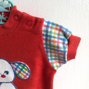 Puede incluir: Camiseta roja de manga corta con una aplicaci&oacute;n de cachorro blanco y ribetes de cuadros en las mangas.