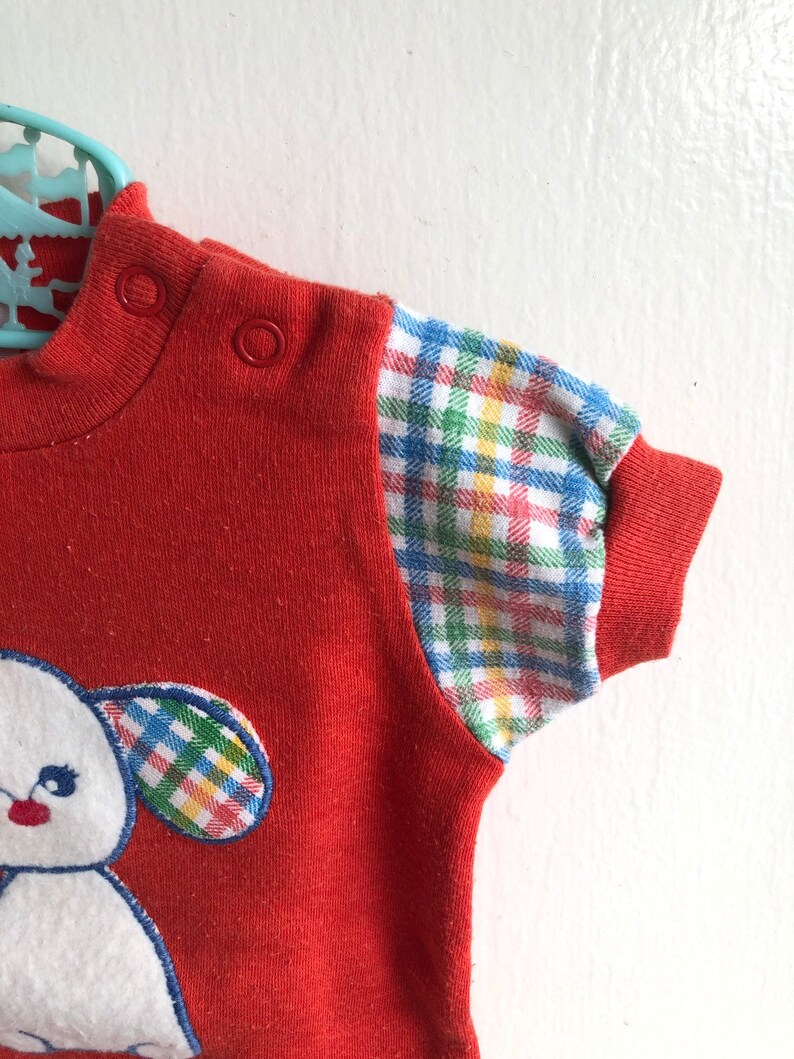 Puede incluir: Camiseta roja de manga corta con una aplicaci&oacute;n de cachorro blanco y ribetes de cuadros en las mangas.