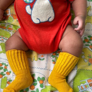 Puede incluir: Un beb&eacute; con un mono rojo con un cachorro blanco bordado en la parte delantera. El beb&eacute; lleva calcetines amarillos y est&aacute; sentado en una manta floral.
