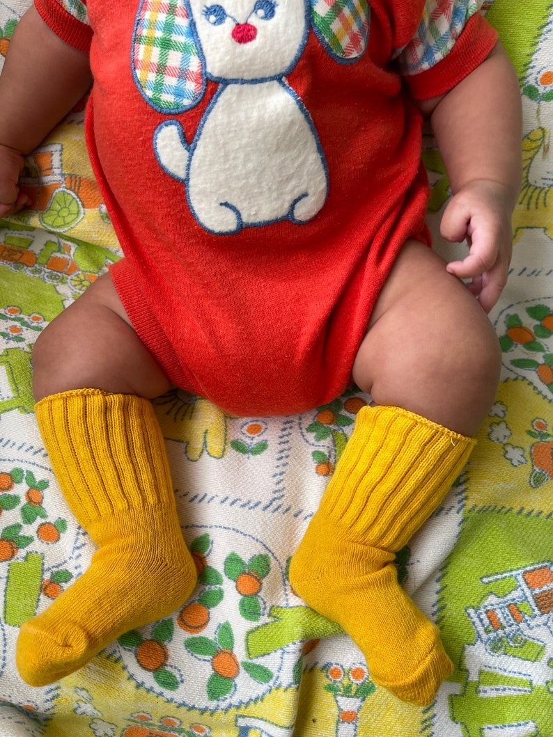 Puede incluir: Un beb&eacute; con un mono rojo con un cachorro blanco bordado en la parte delantera. El beb&eacute; lleva calcetines amarillos y est&aacute; sentado en una manta floral.