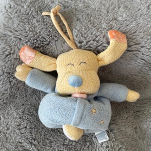 May include: A plush toy dog with a yellow knit head and ears, blue body, and orange accents. The dog is wearing a blue coat with star details. A tag with the brand name "Carter's" is visible.