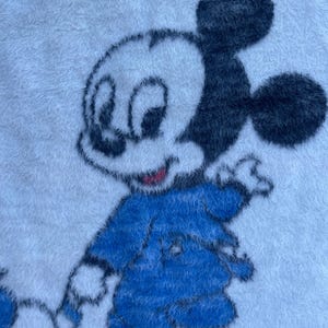 Puede incluir: Toalla de felpa blanca con una imagen de dibujos animados de Mickey Mouse con un atuendo azul. Mickey Mouse tiene orejas negras, una cara blanca y una boca roja. La toalla tiene un aspecto suave y texturizado.