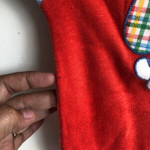 Puede incluir: Mono de beb&eacute; de manga larga rojo con un parche de cuadros en el hombro derecho y una aplicaci&oacute;n de cuadros en la parte delantera. El mono tiene un ribete blanco alrededor del cuello y las mangas.