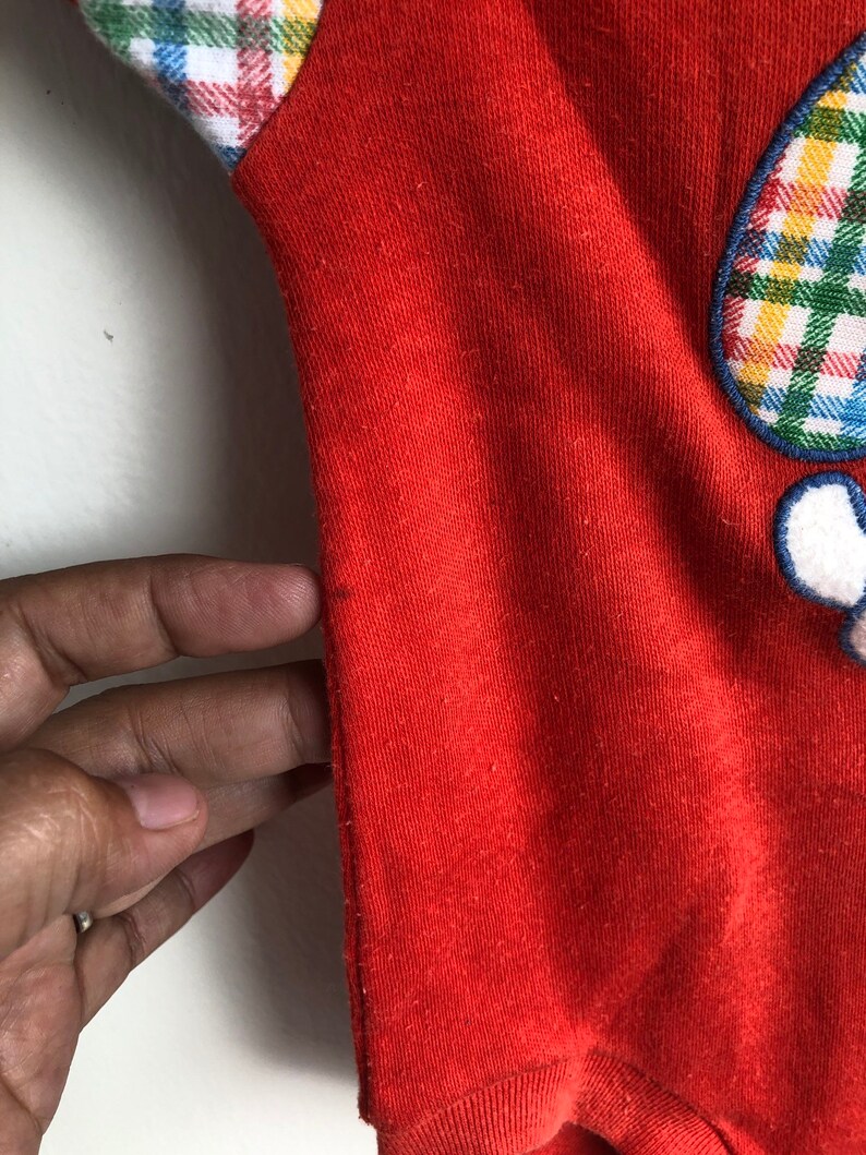 Puede incluir: Mono de beb&eacute; de manga larga rojo con un parche de cuadros en el hombro derecho y una aplicaci&oacute;n de cuadros en la parte delantera. El mono tiene un ribete blanco alrededor del cuello y las mangas.