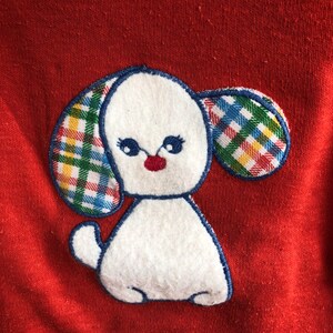 Puede incluir: Sudadera roja con una aplicaci&oacute;n de fieltro blanco de un perro con orejas a cuadros. El perro tiene la nariz roja y los ojos negros.