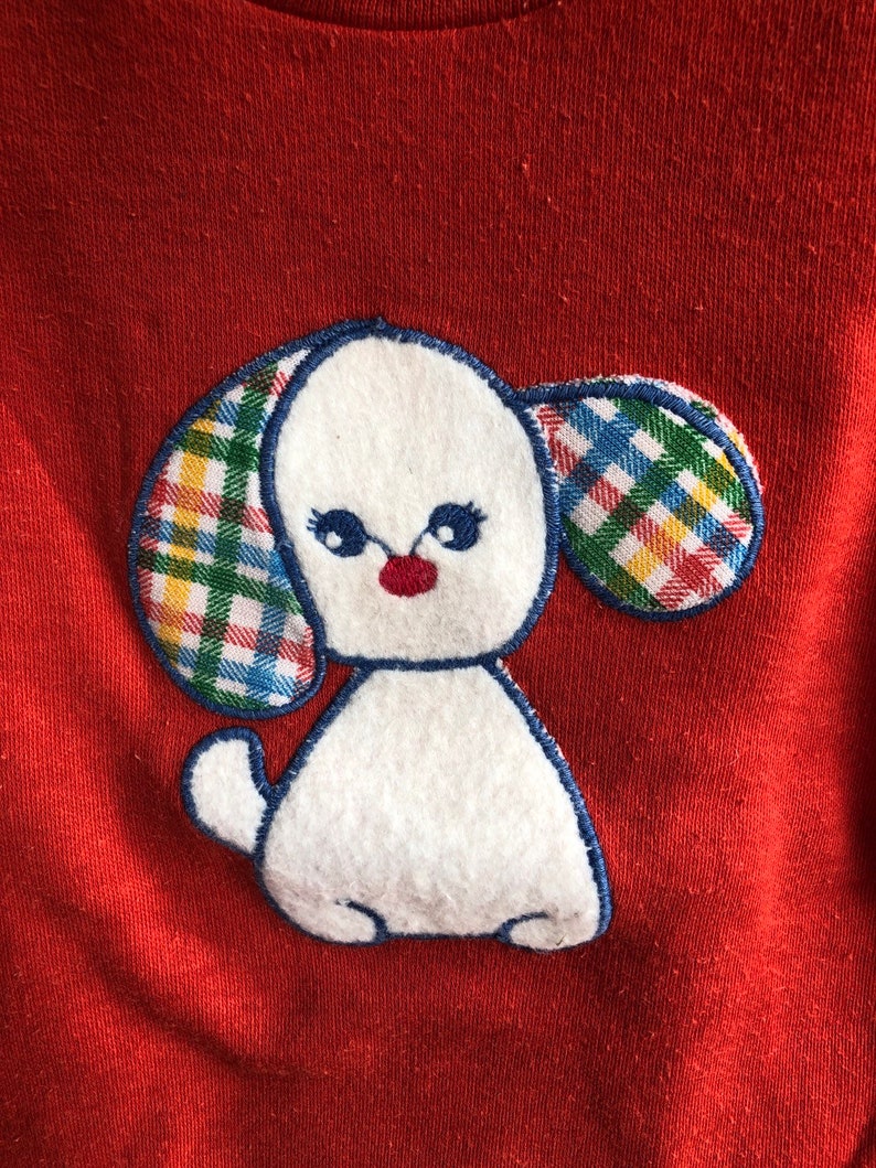 Puede incluir: Sudadera roja con una aplicaci&oacute;n de fieltro blanco de un perro con orejas a cuadros. El perro tiene la nariz roja y los ojos negros.