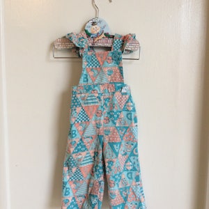 Op de afbeelding: Een turquoise en perzikkleurige patchwork overall met een fantasierijke print van dieren en letters. De overall heeft verstelbare banden en knoopsluitingen.