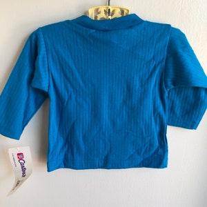 May include: A teal blue long-sleeved shirt with ribbed fabric. The shirt has a tag attached that says "Carter's" and "Made in USA".
