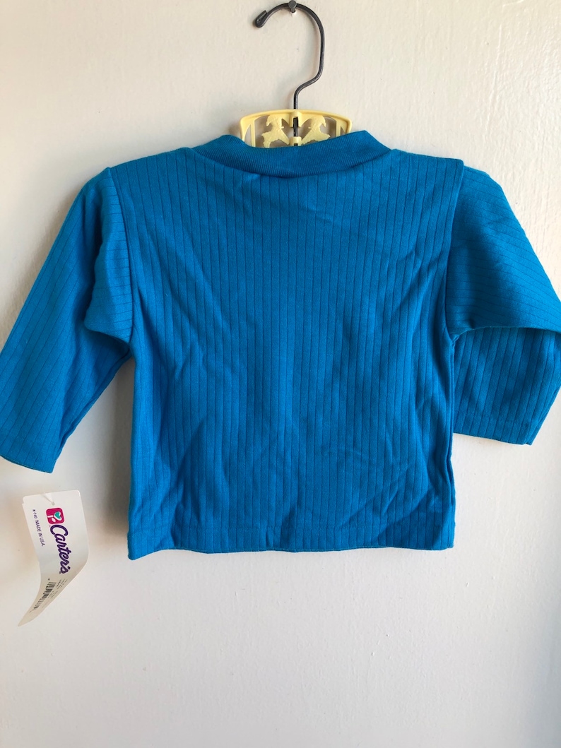 May include: A teal blue long-sleeved shirt with ribbed fabric. The shirt has a tag attached that says "Carter's" and "Made in USA".