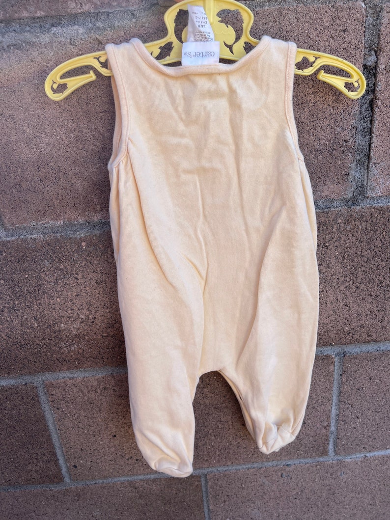 K&ouml;nnte beinhalten: Ein hellgelber, &auml;rmelloser Baby-Strampler mit dem Aufdruck "Carter's" auf der Vorderseite.