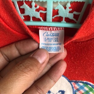 Puede incluir: Un mameluco rojo para beb&eacute; de la marca Carter's con un oso blanco bordado que lleva un lazo de cuadros. El mameluco tiene una etiqueta azul con el texto "Carter's 12 Months 19-22 LBS. 50% Polyester 50% Cotton Machine Wash and Dry Warm Setting No Colors Separately Iron if Needed."