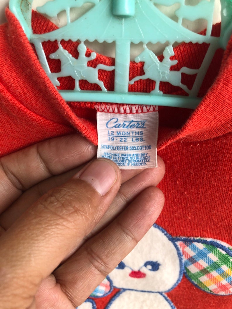 Puede incluir: Un mameluco rojo para beb&eacute; de la marca Carter's con un oso blanco bordado que lleva un lazo de cuadros. El mameluco tiene una etiqueta azul con el texto "Carter's 12 Months 19-22 LBS. 50% Polyester 50% Cotton Machine Wash and Dry Warm Setting No Colors Separately Iron if Needed."